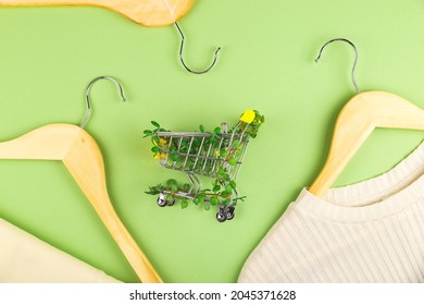 Conscious Consumption Slow Fashion Zero Waste Concept. Shopping Cart Entwined With Plants On Green Background Among Hangers. Sustainable Eco Lifestyle. Top View Flat Lay