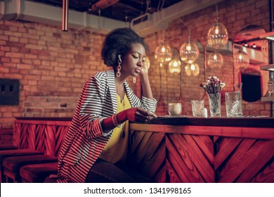 Considering Menu At Bar. Stylish Sophisticated Curly-haired Afro-American Young Female Wearing Striped Cardigan Sitting With Coffee And Looking At Menu At Bar