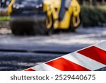 A construction area is marked with bright barriers, while a yellow roller is seen working on the roadway in an urban setting during daylight hours.