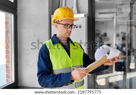 construction business and building concept - male electrician or worker in helmet and safety west with papers on clipboard and pencil at electric board