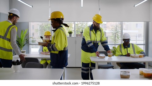 Construction Company Workers Having Takeaway Lunch In Modern Office. Multiethnic Team Of Engineers In Safety Uniform And Hardhat Taking Food From Table In Office