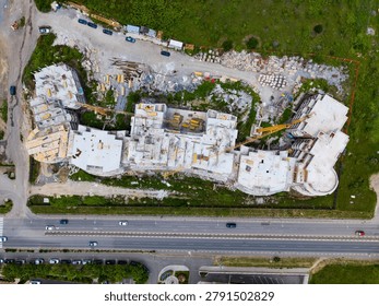 Construction continues at a large site where the building structure is taking shape. Heavy machinery is seen alongside scattered materials. Nearby, cars travel along the road.