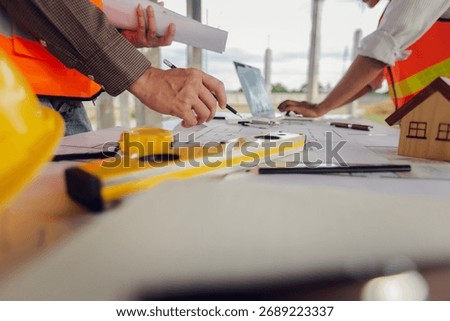 Construction contractor discussing, checking and recording construction work using computer, tape measure, spirit level and wearing yellow construction equipment. House construction concept.