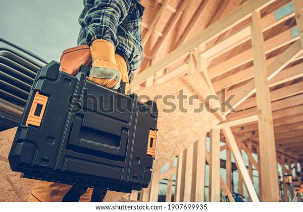 Construction Contractor with Plastic Power Tools\
Box in His Hand Going to Wooden Skeleton Frame Construction Site of\
House. Industrial\
Theme.