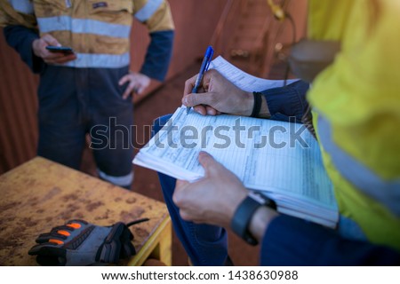 Construction co-worker checking site emergency phone number on cell phone while supervisor whitening on working at height permit prior to performing high risk work construction site open field