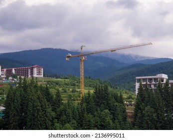 Construction crane among the mountains. Tower crane works on the construction site against the backdrop of a grey cloudy sky and green mountains. Forest encroachment city concept.