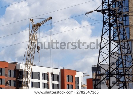 A construction crane is lifting materials close to apartment buildings while power lines run overhead and clouds are visible in the sky.