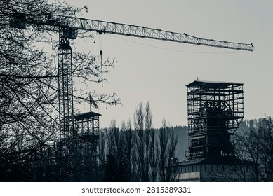 Construction crane and steel tower structure at an industrial site, seen through bare trees under a gray sky