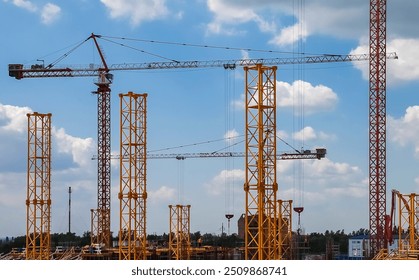 Construction cranes and building frameworks under a bright blue sky in an urban development area. Tall yellow and red cranes tower over the city skyline as workers construct new buildings.