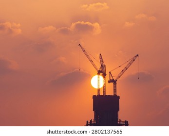 Construction cranes silhouetted against dramatic sunset sky with sun behind building tower structure during golden hour.