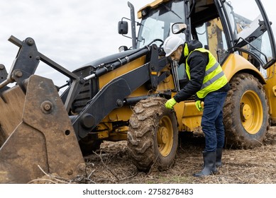 A construction engineer bends beside a backhoe loader to inspect the muddy front wheel and equipment before operation.