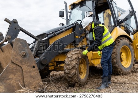 A construction engineer bends beside a backhoe loader to inspect the muddy front wheel and equipment before operation.
