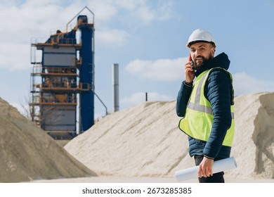 Construction engineer holding blueprints while using a smartphone at a concrete production plant, overseeing the industrial site operations