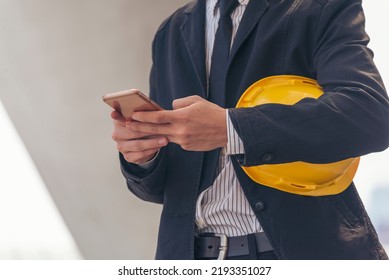 Construction Engineer Holding Yellow Hard Hat Holding Smartphone. Safety Helmet Holding Yellow Hardhat Using Smartphone To Communicate On Construction Site. Hardhat Protect Civil Construction Concept