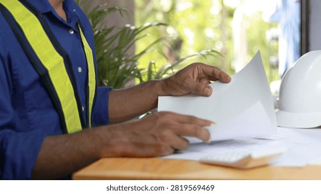 Construction engineer reviewing architectural documents at office desk with reflective safety vest and white hard hat. Engineering planning, project management, workplace safety, building inspection and contractor concept.