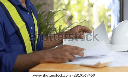Construction engineer reviewing architectural documents at office desk with reflective safety vest and white hard hat. Engineering planning, project management, workplace safety, building inspection and contractor concept.