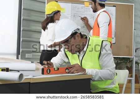 Construction engineer using level tool for precision on desk while team discusses technical drawing on whiteboard in background.