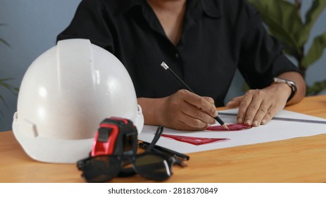 Construction engineer using a pencil and ruler to create technical plans beside a white hard hat, measuring tape, and safety glasses, representing architecture, engineering, design, and workplace safety.
