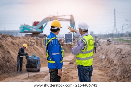 Construction engineer wear safety uniform under inspection and survey workplace by tablet with excavation truck digging and worker construction road background.