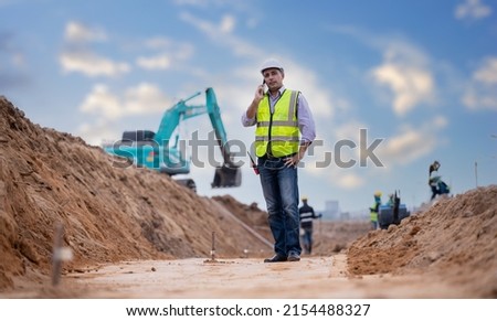 Construction engineer wear safety uniform under inspection and survey workplace by radio communication ,smart phone with excavation truck digging, theodolite and worker construction road background.