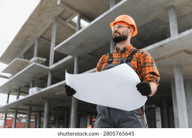 Construction engineer wearing hard hat and safety glasses, holding a blueprint, looking forward while planning on site