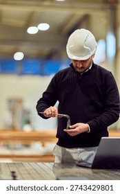 Construction engineer in a workshop inspects a metal part with pliers while wearing a hard hat and smart-casual clothing, laptop nearby. Image conveys focus, professionalism, quality control and workplace safety.