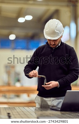 Construction engineer in a workshop inspects a metal part with pliers while wearing a hard hat and smart-casual clothing, laptop nearby. Image conveys focus, professionalism, quality control and workplace safety.