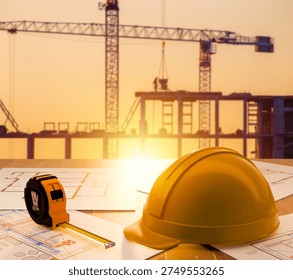 Construction engineering, building development. Safety helmet and measuring tape on architectural blueprints with tower cranes and unfinished building structure at sunset.