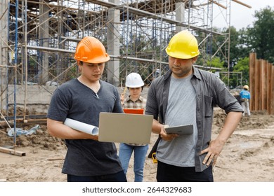 Construction engineers using laptop and tablet to discuss project progress at building site. Team collaboration in civil engineering and site supervision concept.