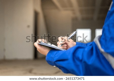 A construction foreman is checking on building quality checklist report during inspecting at the house construction site (as background). Industrial working scene, close-up with selective focus.