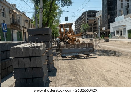 Construction grader machine levels unpaved road surface, preparing for asphalt paving. Concrete curbstones stacked nearby. Urban environment, buildings, infrastructure upgrade.