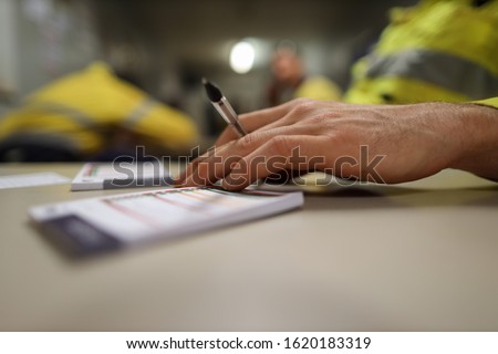 Construction high risk workers planer holding black pen and placing his hand on top of personal safety risk assessment take five booklet on the table with defocused co-teamwork at the background  