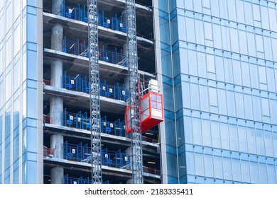 Construction Hoist Lifting. Erection Of High-rise Buildings. Red Temporary Elevator Outside The Skyscraper. Glass High-riser Under Construction. Hoist Lifting A Load At A Construction Site.