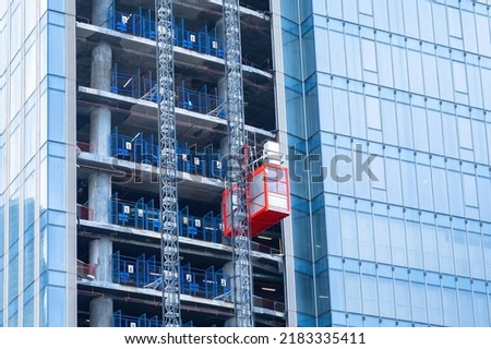 Construction hoist lifting. Erection of high-rise buildings. Red temporary elevator outside the skyscraper. Glass high-riser under construction. Hoist lifting a load at a construction site.