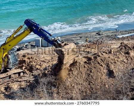 A construction machine is digging in the dirt near the beach. Workers are present on the site, and the ocean waves can be seen in the background. The weather is clear and bright.