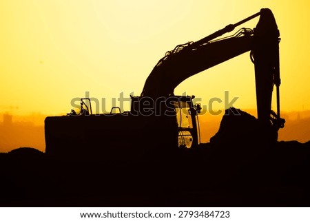A construction machine operates on a worksite at sunset. It moves dirt as the sun sets behind the horizon, casting a silhouette against the sky