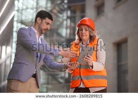 Construction manager and engineer discussing a problem at the building site. Female professional looks confused while checking information on a tablet.