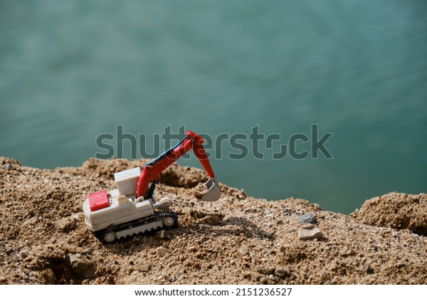Construction and mining industry. Creative banner.\
Small plastic toy excavator with bucket working on sand extraction\
at quarry. Pond in background. Children\'s toy model of tractor\
extracts sand.