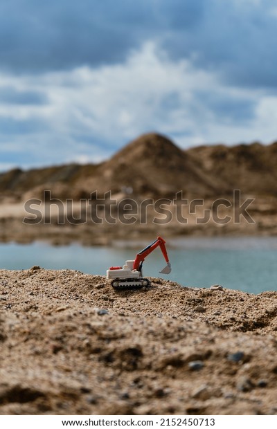 Construction and mining industry. Creative banner.\
Small plastic toy excavator with bucket working on sand extraction\
at quarry. Pond in background. Children\'s toy model of tractor\
extracts sand.