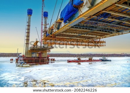 Construction of a new road bridge continues across the Sheksna river. Cable-stayed bridge in Cherepovets at sunset. Fragment of the unfinished construction of the city highway.Cargo barge on ice. 