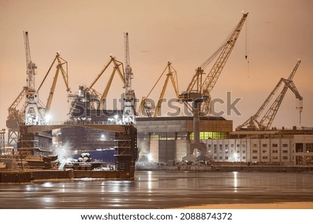 The construction of nuclear icebreakers at night, cranes of of the Baltic shipyard in a frosty winter day, steam over the Neva river, smooth surface of the river, fog
