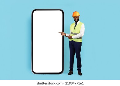 Construction Offer. Smiling Black Male Engineer Pointing At Huge Blank Smartphone With White Screen, African American Civil Engineer In Safety Vest And Hard Hat Recommending New Mobile App, Mockup