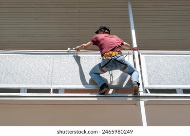 A construction painter paints a balcony while hanging from a safety harness on the balcony of a high-rise building during daylight hours. - Powered by Shutterstock