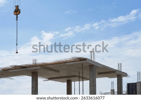 Construction process of a concrete building with exposed rebar and supporting columns against a bright blue sky. A crane hook for construction work is suspended above