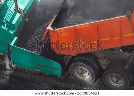 construction process where an orange dump truck is tipping fresh, hot asphalt mix into the hopper of a green asphalt paver machine, illustrating road construction and infrastructure work