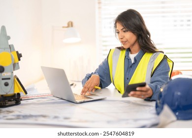 Construction professional reviewing plans on laptop in office with tools and safety gear present during work hours - Powered by Shutterstock