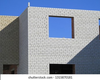 Construction progress: partially built structure showcases light-colored brickwork with uniform pattern, reflecting skilled masonry craftsmanship. Two rectangular window openings frame vibrant blue sky, enhancing symmetrical facade design while revealing internal framing materials that indicate ongoing building efforts. Natural sunlight casts soft shadows across wall, creating depth through clear distinction of light and dark areas. This scene communicates development potential and anticipation, suitable for real estate marketing, architecture industries, and urban construction themes. Image focuses on exterior with sky backdrop, providing scale and perspective for commercial use. Clarity of details and lighting highlights structural aspects, appealing for design element imagery.