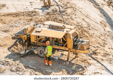 Construction scene featuring mobile drilling rig, worker inspecting equipment, high-visibility safety gear, loose soil, rocks, uneven terrain, daytime lighting, bright yellow machine, earthy tones
