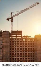 A construction site is bustling with activity as workers are visible at various levels of the structure. A crane towers above, silhouetted against the vibrant sunset sky.