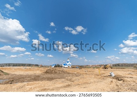 Construction site with cleared land and a distant blue and white church under a bright blue sky with scattered clouds in a rural area